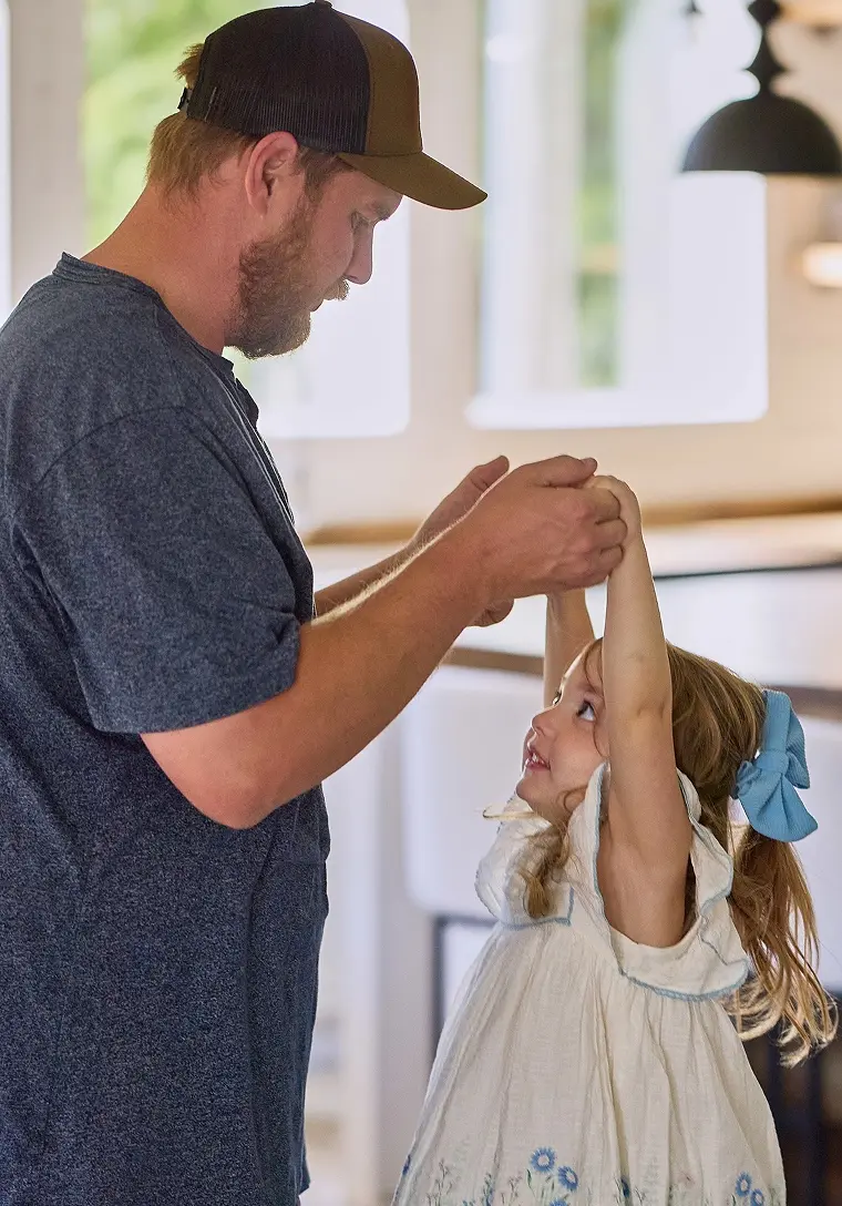 A father dances with his young daughter, holding her hands up, inside their home.
